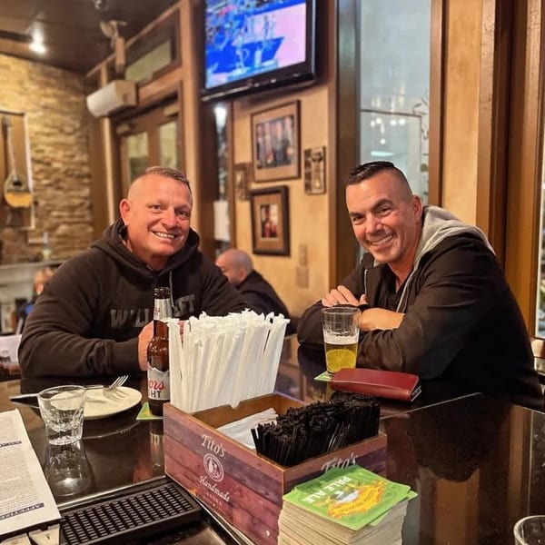 Two smiling men sit at a bar with drinks, enjoying the lively atmosphere of Demitris Taverna.