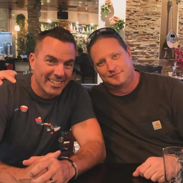 Two men smiling at the camera in Demitris Taverna, with Greek decor in the background.