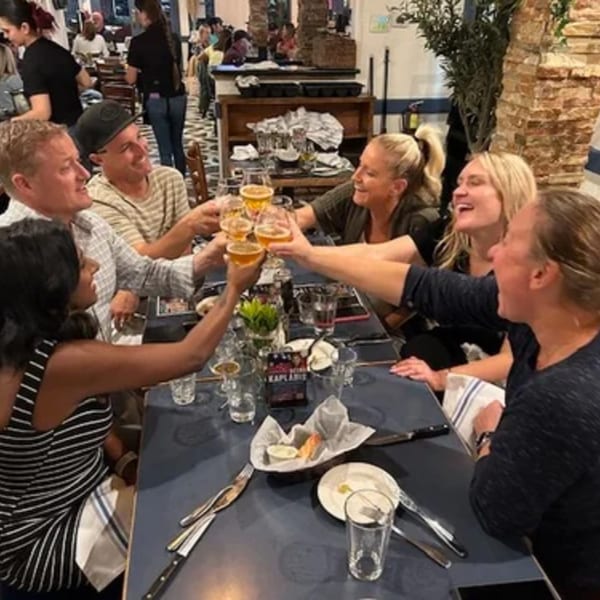 Friends at Demitris Taverna clinking glasses of beer, laughing, and enjoying their meal.
