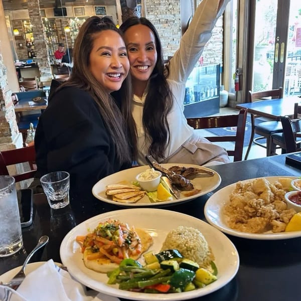 Two smiling women at Demitris Taverna with plates of Greek food, including calamari and kebabs.
