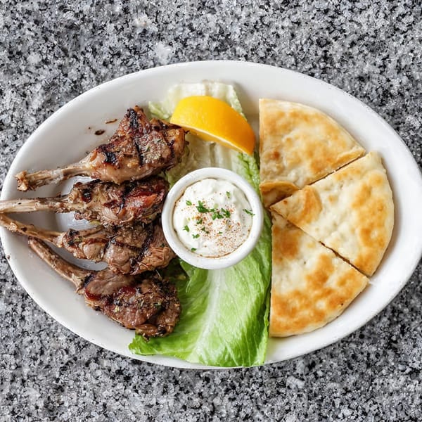 Grilled lamb chops, pita bread, tzatziki sauce, lettuce, and a lemon wedge on a white plate.