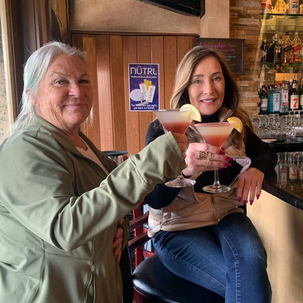 Two women at Demitris Taverna bar, smiling and toasting pink cocktails with lemon garnishes.